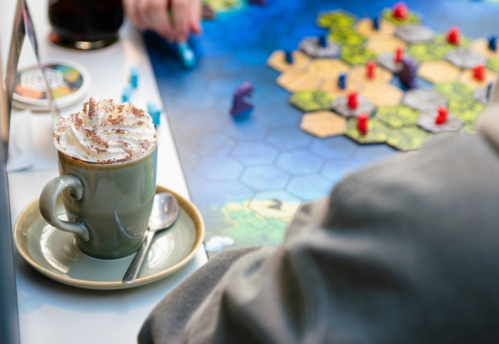 A close-up of a cappuccino sitting on a table, next to a board game. A hand is moving one of the pieces from the game. 
