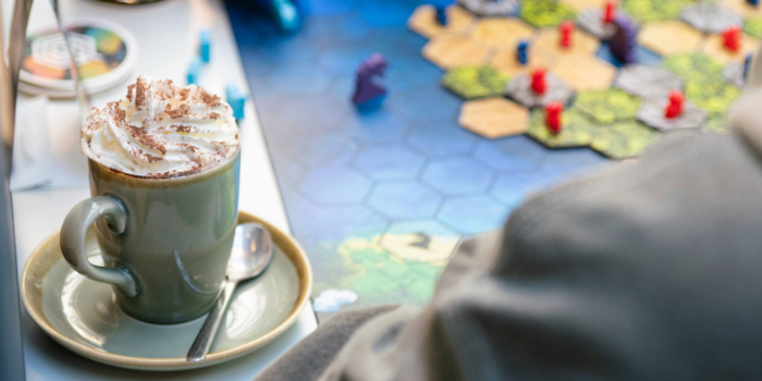A close-up of a cappuccino sitting on a table, next to a board game. A hand is moving one of the pieces from the game. 