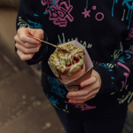 Close-up of someone holding a dessert covered in toppings and sauce in a cardboard holder.