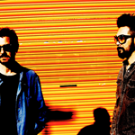 A picture of the band, which consists of two individuals, who are both wearing sunglasses, and stood infront of an orange shutter