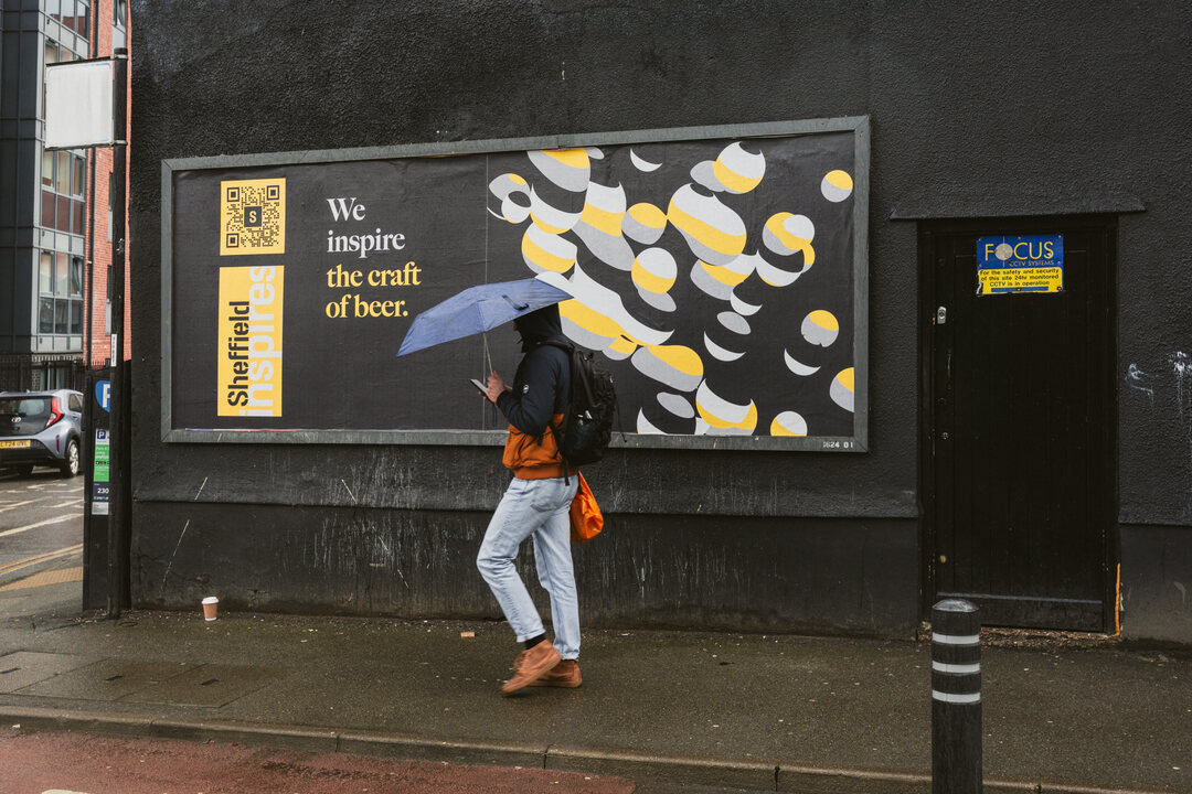 A person walking along a city street in the rain, holding an umbrella and passing a large billboard that reads “We inspire the craft of beer,” featuring bold graphic artwork on a dark wall.