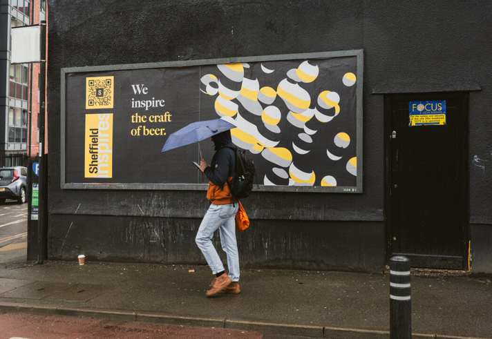 A person walking along a city street in the rain, holding an umbrella and passing a large billboard that reads “We inspire the craft of beer,” featuring bold graphic artwork on a dark wall.