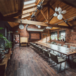 Inside The Chimney House . Spacious meeting room with exposed brick walls, wooden ceiling beams, and large skylights allowing natural light. A long rectangular table with modern chairs runs down the center of the room. Pendant lights and spotlights hang from the ceiling, and plants decorate the side tables and windowsills. A large wall clock and framed board are mounted on the far wall, adding to the rustic-industrial design.