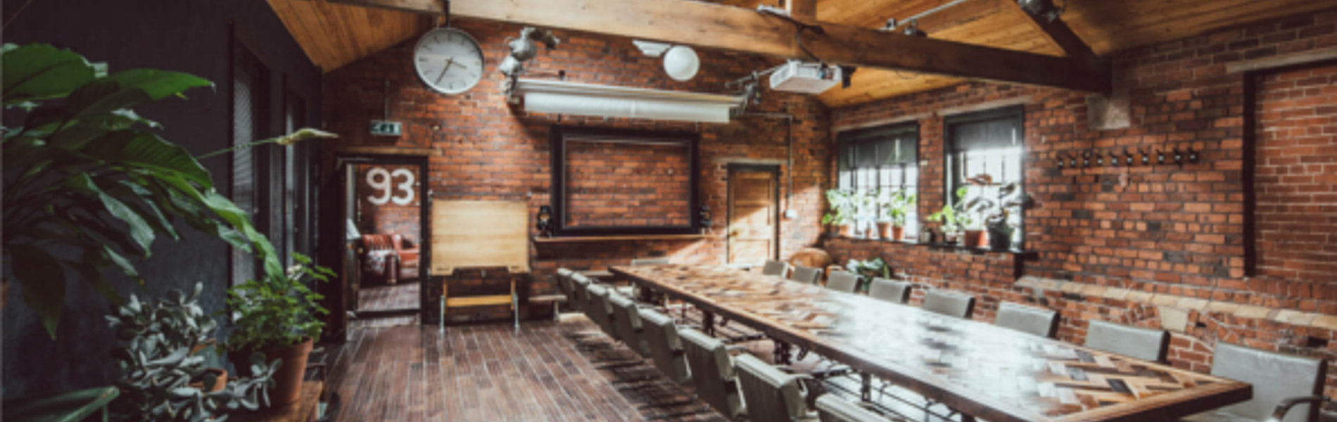 Inside The Chimney House . Spacious meeting room with exposed brick walls, wooden ceiling beams, and large skylights allowing natural light. A long rectangular table with modern chairs runs down the center of the room. Pendant lights and spotlights hang from the ceiling, and plants decorate the side tables and windowsills. A large wall clock and framed board are mounted on the far wall, adding to the rustic-industrial design.