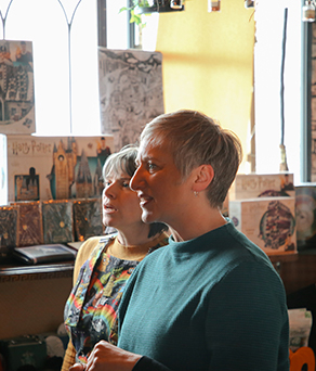 Two women look at various artworks on the walls of a studio.