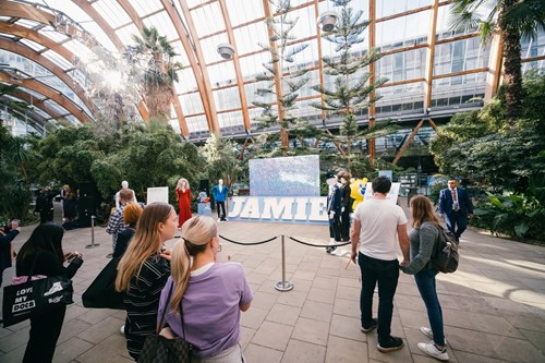 A group of people gather inside a spacious, glass-roofed conservatory filled with lush greenery and tall trees. A large sign reading 'JAMIE' stands in the center. Some attendees take photos while others explore the surroundings, with natural light streaming through the glass ceiling.