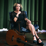 A person seated in a patterned armchair with legs crossed, wearing a black dress and black shoes. The setting includes a dark green curtain backdrop and a polished floor with a mix of colours. A wooden acoustic guitar rests on the floor in front of the chair, leaning slightly toward the person.