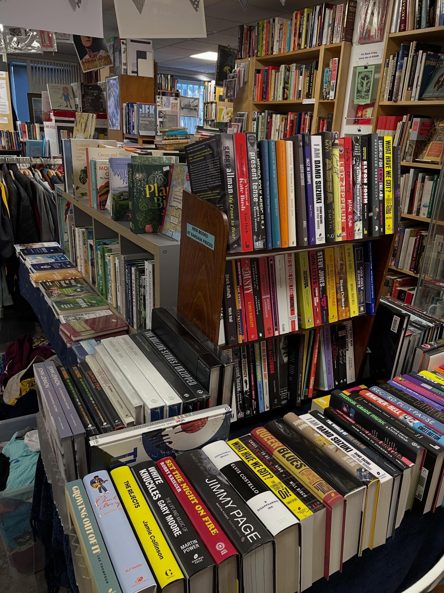 A crowded second-hand bookshop interior with shelves and tables stacked with books of various sizes and colors. Prominent titles include music biographies and art books. The foreground shows a table filled with hardback books arranged in neat rows, while the background has tall shelves packed with books and some clothing racks visible on the left.