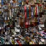 A crowded second-hand bookshop interior with shelves and tables stacked with books of various sizes and colors. Prominent titles include music biographies and art books. The foreground shows a table filled with hardback books arranged in neat rows, while the background has tall shelves packed with books and some clothing racks visible on the left.