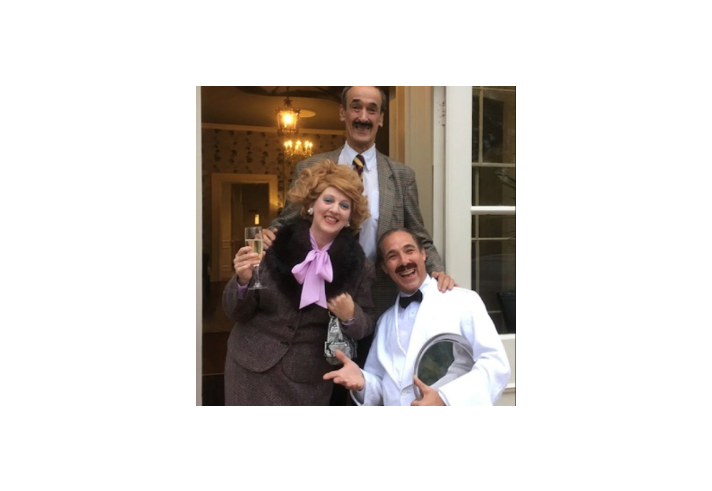 Three people are standing in front of an open doorway with glass panels. The person on the left is wearing a brown textured suit with a large black fur collar and a bright pink bow tie, holding a champagne flute in one hand and a handbag in the other. The person in the middle is dressed in a grey checked blazer with a white shirt and striped tie. The person on the right is wearing a white tuxedo jacket with a black bow tie and is holding a silver serving tray. 