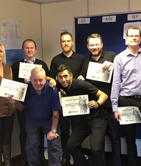 A dozen people standing in a workspace. They are all holding certificates and smiling at the camera.