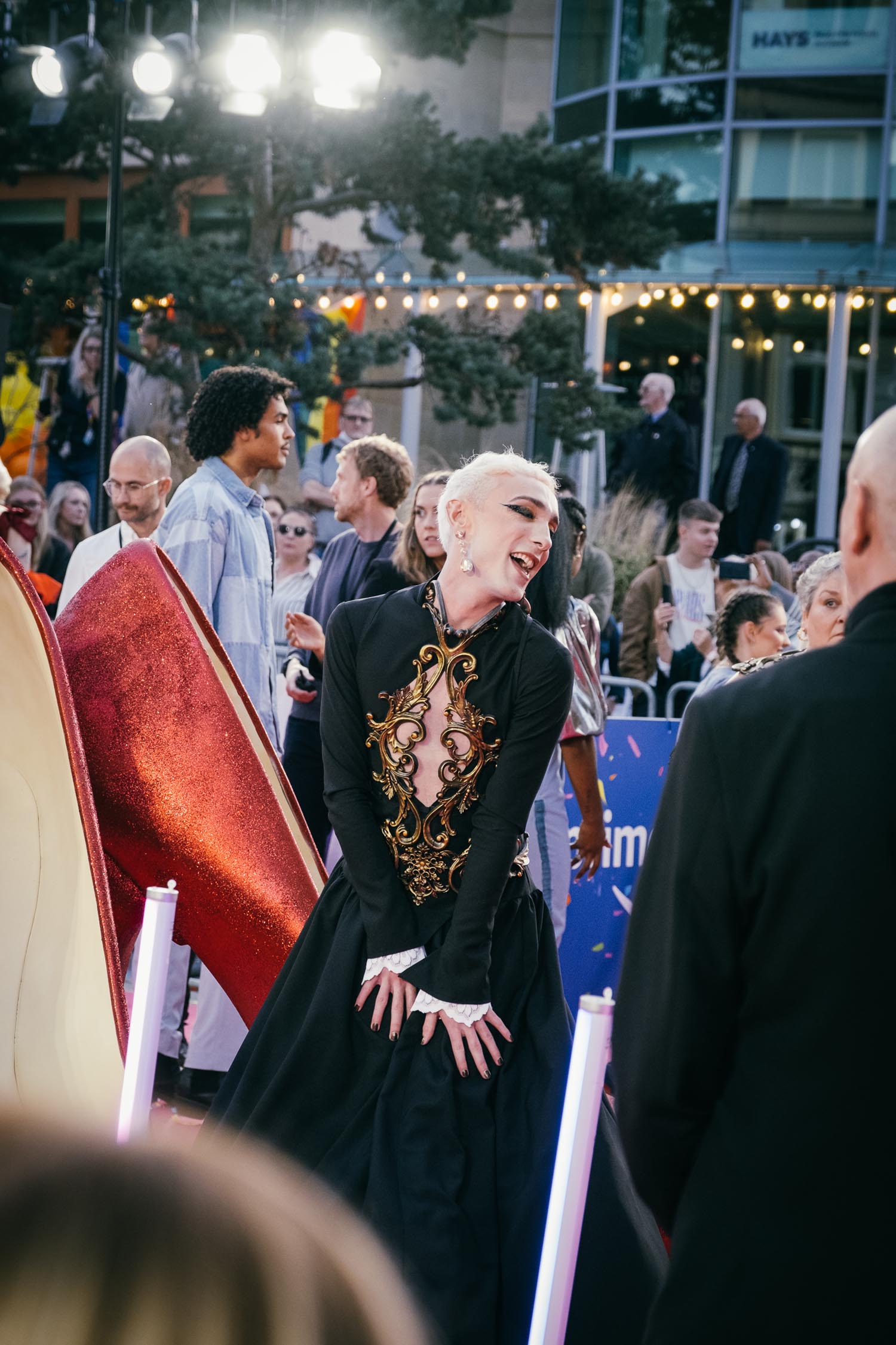 A person in an elaborate black outfit with gold detailing poses on a red carpet. In the background, a crowd watches and takes photos under festive string lights. Large decorative high-heeled shoe props are visible in the foreground.
