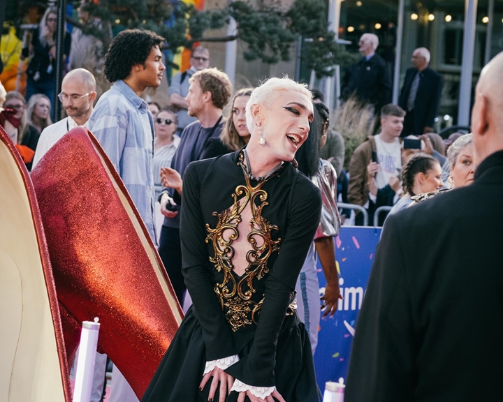 A person in an elaborate black outfit with gold detailing poses on a red carpet. In the background, a crowd watches and takes photos under festive string lights. Large decorative high-heeled shoe props are visible in the foreground.
