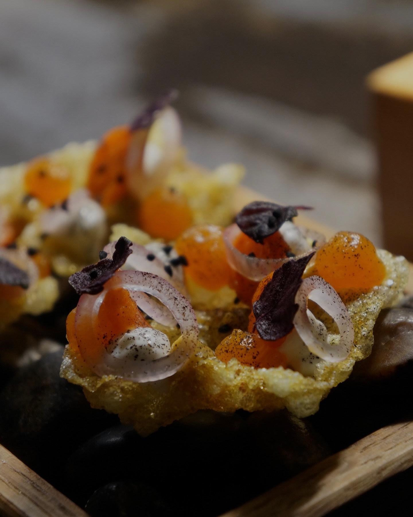  Close-up of an intricate dish featuring crisp golden wafers topped with orange spheres, thinly sliced shallots, and purple herb leaves, arranged on a wooden tray with dark stones beneath.