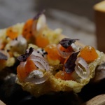  Close-up of an intricate dish featuring crisp golden wafers topped with orange spheres, thinly sliced shallots, and purple herb leaves, arranged on a wooden tray with dark stones beneath.