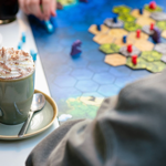 A close-up of a cappuccino sitting on a table, next to a board game. A hand is moving one of the pieces from the game. 