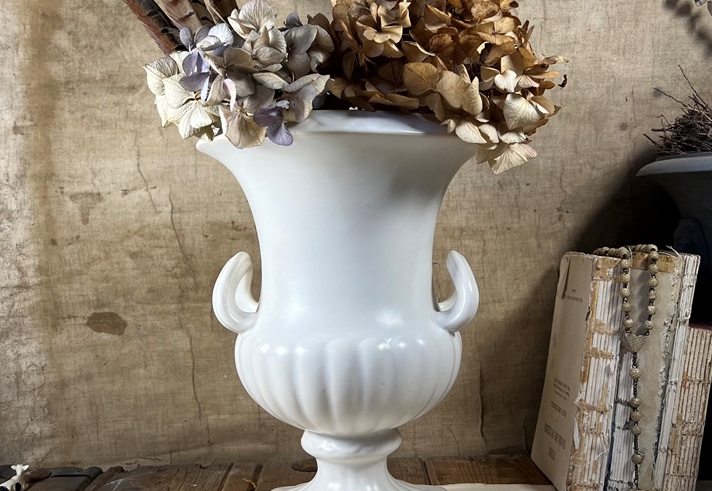 A white porcelain vase, in the style of a Greek urn, with dried flowers in.