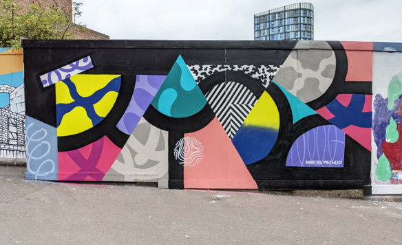 Street art mural featuring bold geometric shapes and patterns in vibrant colors including teal, pink, yellow, blue, and black. The design includes triangles, circles, and abstract textures on a black background, painted on an outdoor wall in an urban setting with a tall modern building visible in the background.