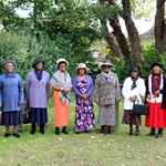 A group of ladies of African Heritage stand in a row in a garden or a park.