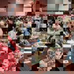 A busy indoor pub or event hall where groups of people sit around tables participating in what appears to be a quiz night. In the foreground, several individuals sit at a wooden table covered with drinks, pens, papers, and raffle-style tickets. More tables filled with attendees extend into the background, and the room features red walls, wooden flooring, and soft overhead lighting. A banner is visible near the back of the room, adding to the lively, social atmosphere.