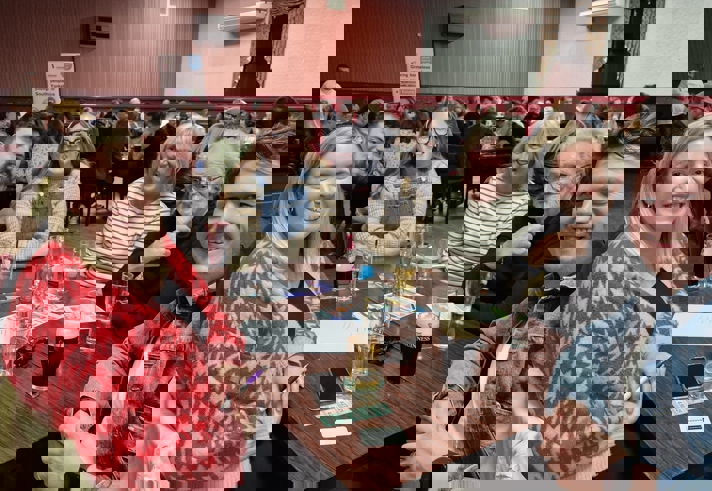 A busy indoor pub or event hall where groups of people sit around tables participating in what appears to be a quiz night. In the foreground, several individuals sit at a wooden table covered with drinks, pens, papers, and raffle-style tickets. More tables filled with attendees extend into the background, and the room features red walls, wooden flooring, and soft overhead lighting. A banner is visible near the back of the room, adding to the lively, social atmosphere.