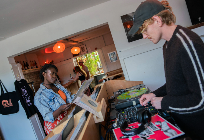 A person stands at a wooden record counter browsing vinyl records, while another operates DJ equipment with turntables and a mixer. Headphones and a colourful record sleeve are visible on the counter. The setting appears to be a cosy shop or café with white walls, hanging orange lights, shelves with bottles, and a tote bag displayed on the wall. A doorway in the background reveals more people and greenery outside.