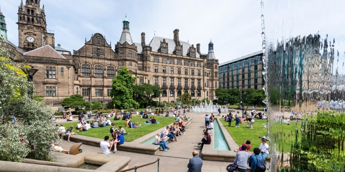 A view of the Peace Gardens on a sunny day. People are walking about while others are sitting on the grass. In the background is Sheffield Town Hall.