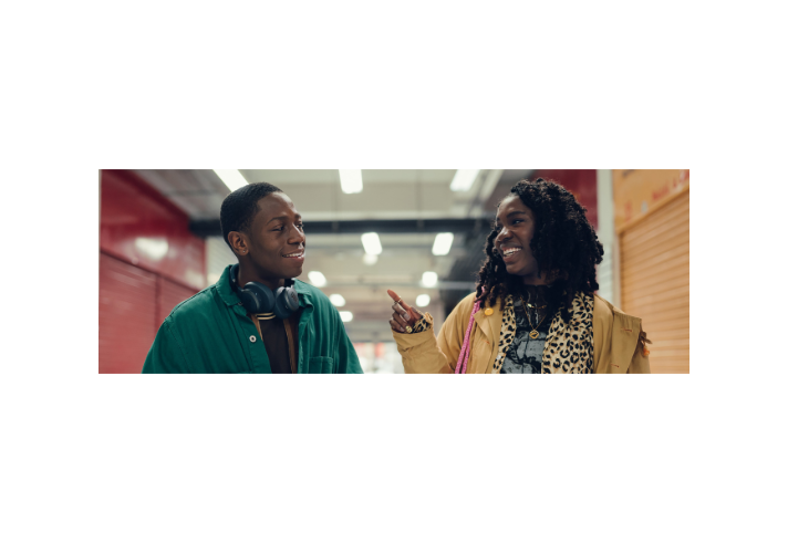 Two people standing in an indoor setting with bright overhead lights and red and orange walls. One person wears a green jacket over a dark top with headphones around the neck. The other person wears a tan coat layered over a leopard-print scarf and graphic T-shirt, accessorised with gold jewellery and a pink strap bag. The second person gestures with one hand while speaking, suggesting a casual conversation in a corridor-like space.