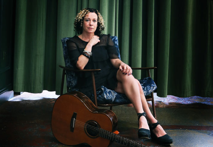 A person seated in a patterned armchair with legs crossed, wearing a black dress and black shoes. The setting includes a dark green curtain backdrop and a polished floor with a mix of colours. A wooden acoustic guitar rests on the floor in front of the chair, leaning slightly toward the person.