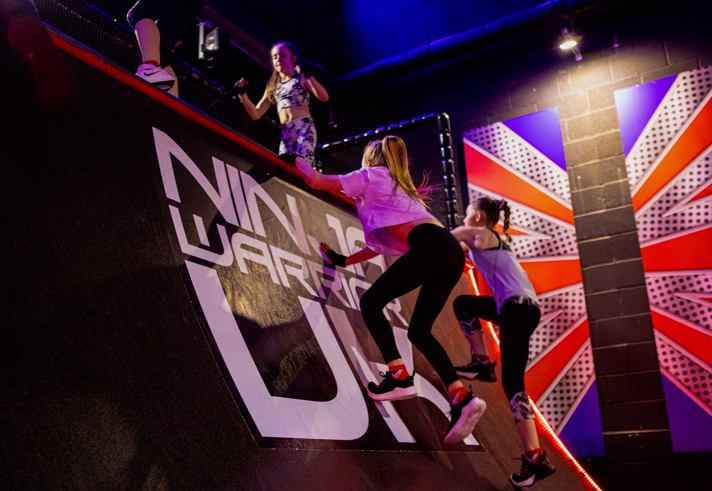 A group of people climb a steep, curved obstacle wall inside an indoor activity venue styled with bright lighting and bold graphics. The wall features large text reading “NINJA WARRIOR UK,” and the surrounding décor includes a vibrant mural with red, blue, and white geometric patterns. Multiple participants are mid-climb, using the wall’s curved surface to reach the top ledge while others stand above the obstacle.