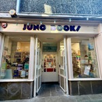 A closer view of the “Juno Books” storefront, showing the cream-painted façade, large display windows, and an open doorway. Colourful posters and book displays are visible through the windows, and fairy lights are strung across the top of the shop.