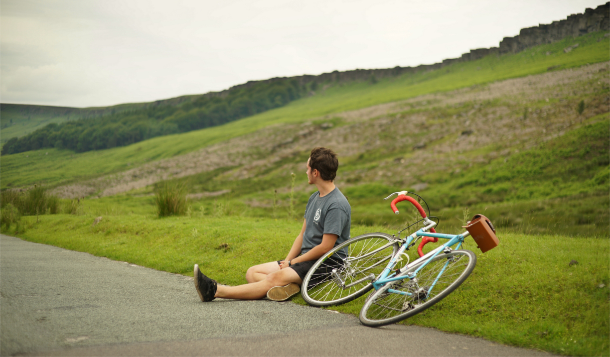 A cyclist sat on the edge of the road having a rest, out in the countryside, with his bike lying on the ground.