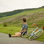 A cyclist sat on the edge of the road having a rest, out in the countryside, with his bike lying on the ground.