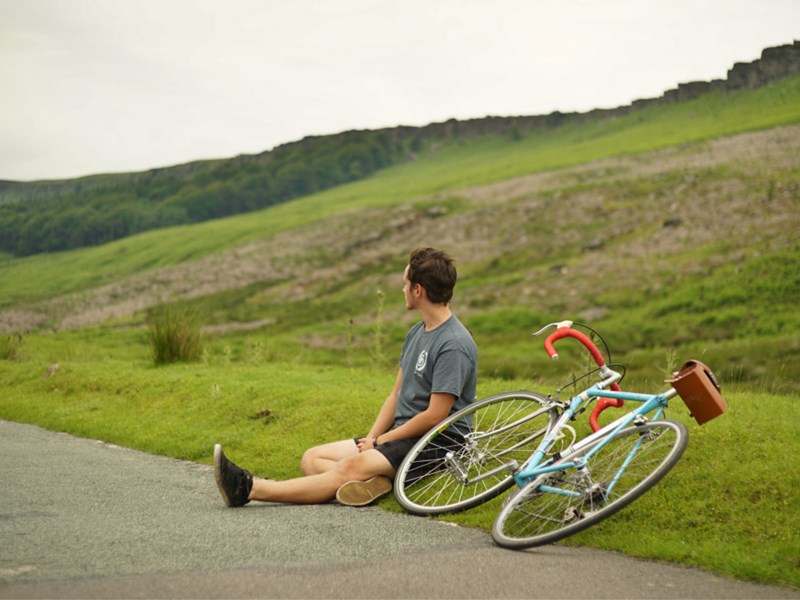 A cyclist sat on the edge of the road having a rest, out in the countryside, with his bike lying on the ground.
