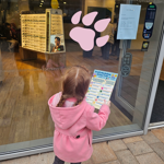 A child stands in front of a shop, with a check list, looking at a large pink paw print on the shop window.
