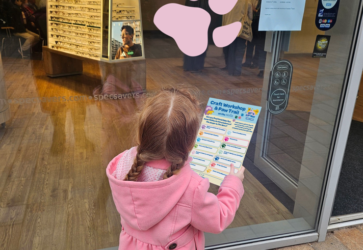 A child stands in front of a shop, with a check list, looking at a large pink paw print on the shop window.
