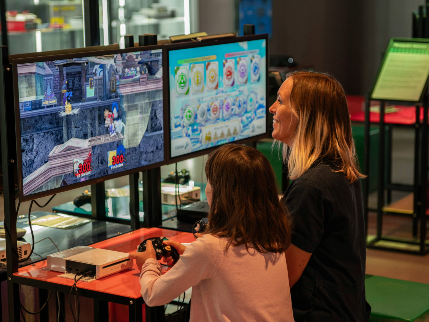 A person with a child sat next to them are playing a video game together. In the background are displays of video games from the history of the video game industry.