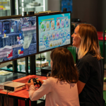 A person with a child sat next to them are playing a video game together. In the background are displays of video games from the history of the video game industry.