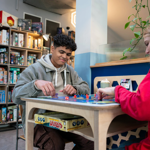 Two people are sat at a table, in a cafe, playing a board game and drinking coffees. Behind them is shelving stacked full of all different board games.