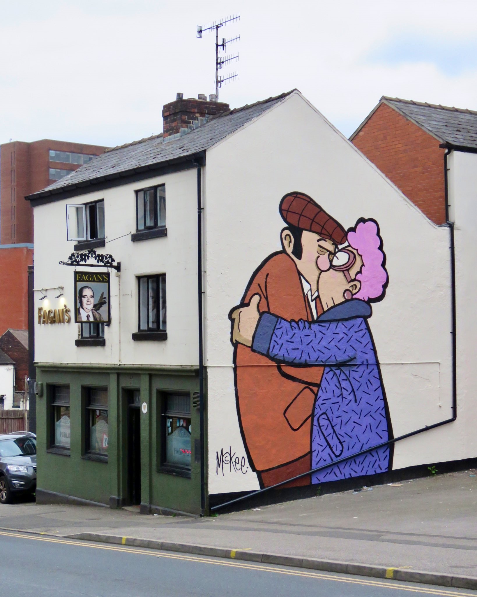 Street art mural on the side of a white building depicting two cartoon-style characters embracing, one wearing an orange coat and flat cap and the other in a purple coat with curly pink hair. The artwork is signed ‘McKee’ and located above a pub called Fagans.