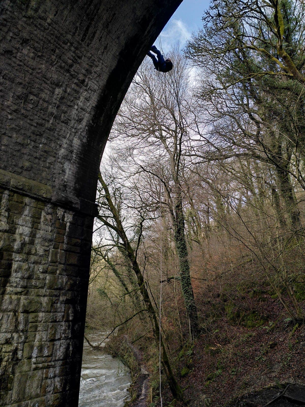 A tall stone arch of a bridge over a river. A person is abseiling down the side.