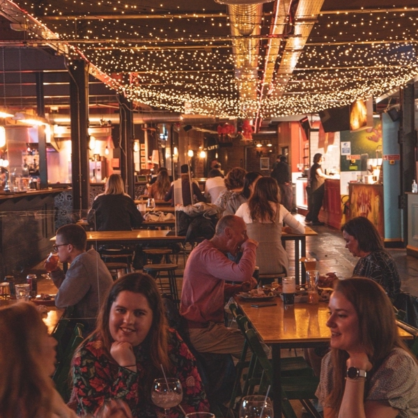 A view inside Cutlery Works: there are people sat at tables eating, chatting and having a good time. The ceiling is festooned with twinkling fairy lights. 