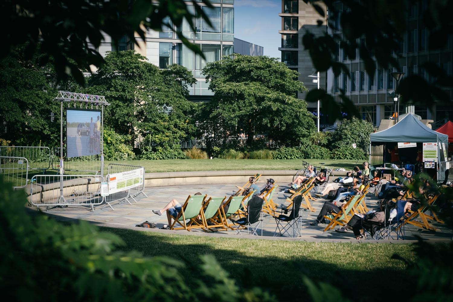 Outdoor screening area with people seated on deck chairs and folding chairs in front of a large screen, surrounded by greenery and modern buildings.