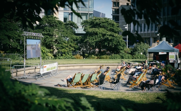 Outdoor screening area with people seated on deck chairs and folding chairs in front of a large screen, surrounded by greenery and modern buildings.
