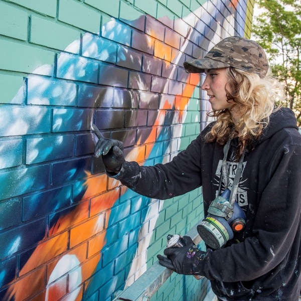 A Person wearing a dark hoodie and cap painting a colorful mural on a green brick wall using spray paint, with shades of blue, orange, and white forming abstract shapes. The scene includes a metal railing and leafy trees in the background.