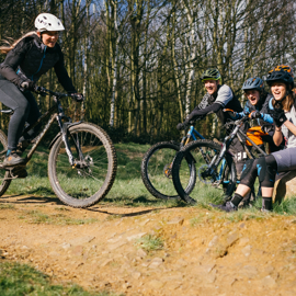 Female mountain biker riding on dirt track. Three other bikers are sat to the right on a log cheering her on.