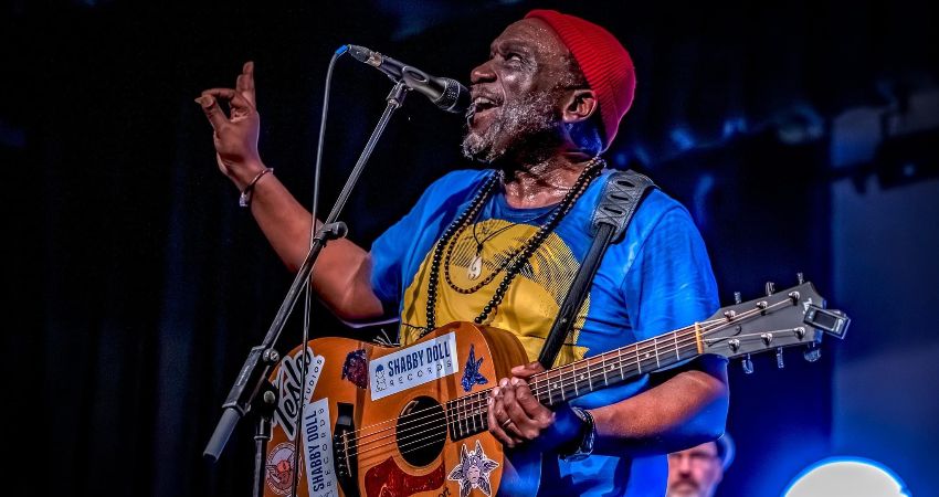 Musician performing on stage wearing a bright blue shirt and red knit hat, playing an acoustic guitar decorated with colorful stickers, including one that reads “Shabby Doll Records.” The performer is standing in front of a microphone, gesturing with one hand while holding the guitar strap over the shoulder. Stage lighting creates a vivid atmosphere with blue and purple tones in the background.