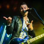 A musician on stage wearing a black leather jacket and white shirt, holding an electric guitar with a shiny blue finish. The performer stands in front of a microphone, gesturing with both hands as colourful stage lights create a vibrant backdrop with green and yellow tones.