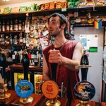 A person behind a bar pulling a pint from a row of hand-pulled beer pumps, with shelves of snacks, bottles, and decorations filling the background.