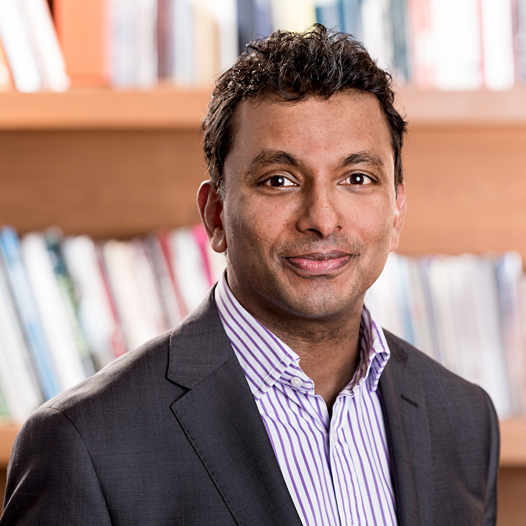 Person wearing a dark suit jacket over a light-colored, striped collared shirt, standing in front of a wooden bookshelf filled with books.